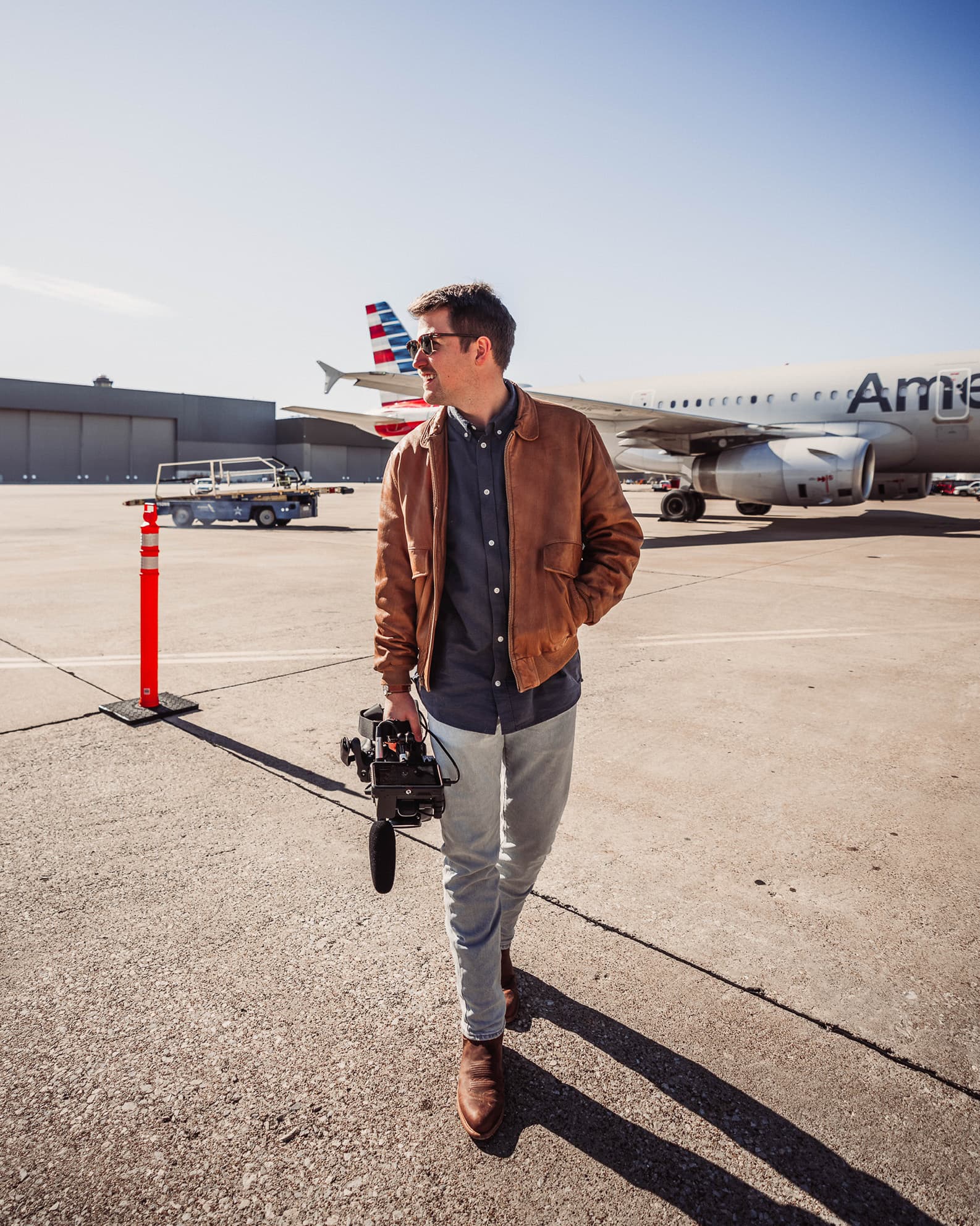 Luke Heslep walking across an airport tarmac with a cinema camera, American Airlines jet behind him