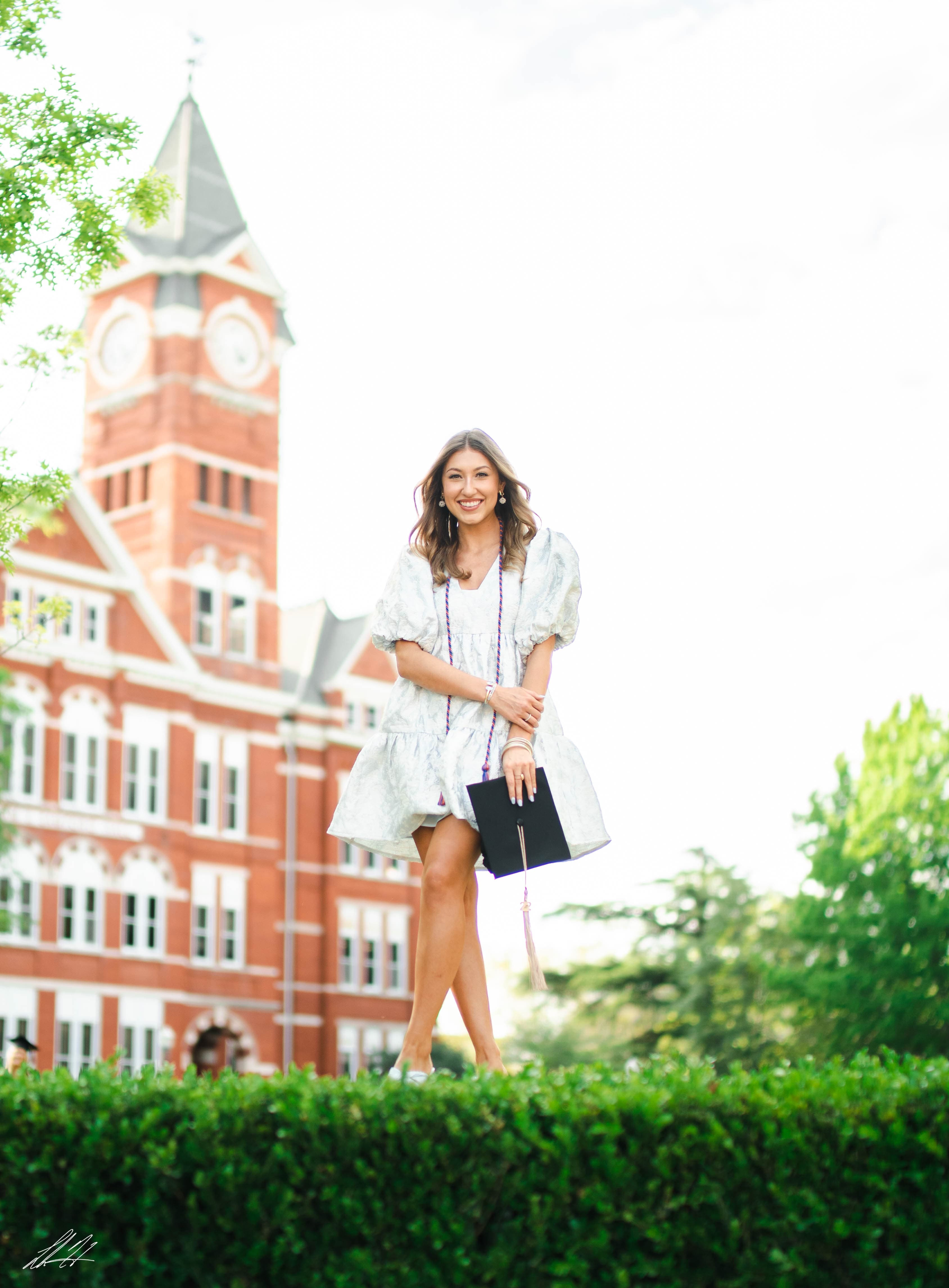 Graduate in a white dress holding a mortarboard on campus